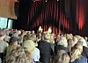 Publikum sitter tett i en sal mens to personer står på en scene foran røde gardiner.